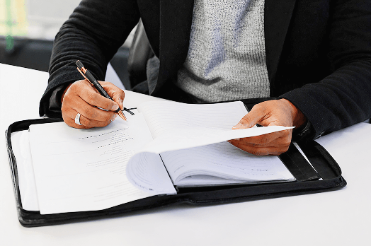 Person reviewing documents and writing notes on a pen with a folder on a white desk.