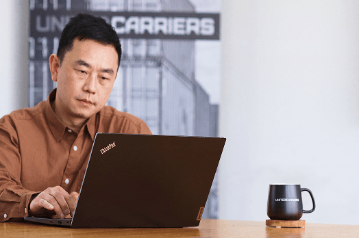 Man in brown shirt working on a ThinkPad laptop at a table with a black mug labeled Brutelo Carriers.